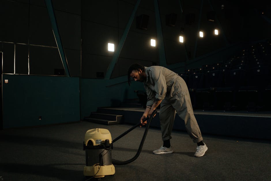 A person performing surface cleaning with a vacuum cleaner, which has a yellow body and black hose, on a patterned area rug in a well-lit room. The individual is holding a white cloth and vacuuming th