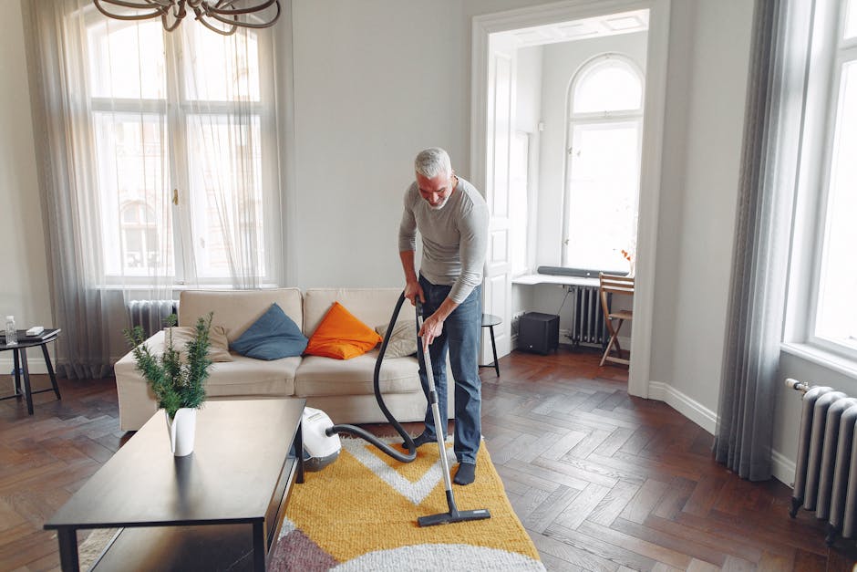 Close-up image showing a person using a red vacuum cleaner on a beige textured carpet in a residential room. The individual's bare feet are visible, standing on the carpet, with part of their black cl