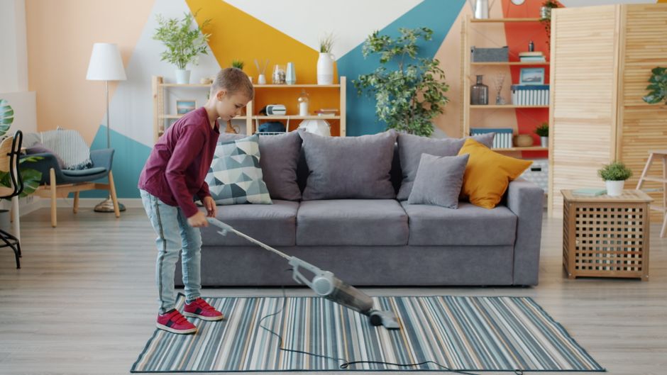 A young boy using a handheld vacuum cleaner to clean a striped area rug in a modern living room with light wooden flooring, a grey fabric sofa with decorative cushions, a wooden side table, and colorf